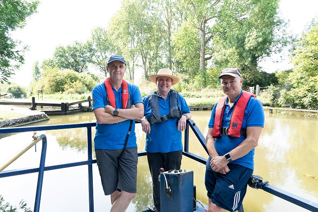Photo of 3 CanalAbility volunteers smiling stasning at the rear of a canal boat with the river behind.