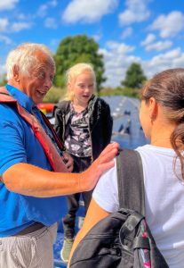 Photo of a CanalAbility volunteer smiling at a lady. There is a young girl and a lady. The girl is in the background smiling towards the camera. The lady is facing away from the camera and you can see her from the back. You can see they are on a canal boat.