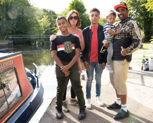 Photo of a family, mum and dad with two teenage boys and the dad is holding a young child in his arms and he has his thumb up. They are standing next to the canal witth the front section of a blue and red canal boat in the foreground.boat in the foreground.
