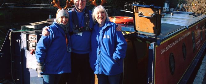 Three people wearing Christmas antlers standing on the back of a canal boat