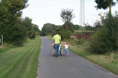 A man pushes a wheelchair while walking with a young girl on a rural road lined with grass and trees.
