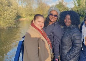 Three ladies are smiling, standing on a canal boat with the river and trees in the background.