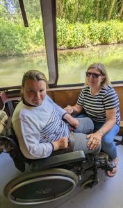 A young wheelchair user is inside a canal boat with another lady kneeling in front of her. The river and greenery can be seen in the background.