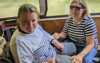 A young wheelchair user is inside a canal boat with another lady kneeling in front of her. The river and greenery can be seen in the background.