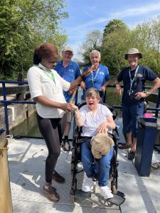 A lady in a wheelchair is smiling and steering a canal boat. Her carer is next to her helping, along with three CanalAbility volunteers.