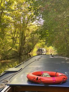 Photo is looking down a canal boat towards the front with a life ring in shot and autumnal trees and a narrowboat in the background.